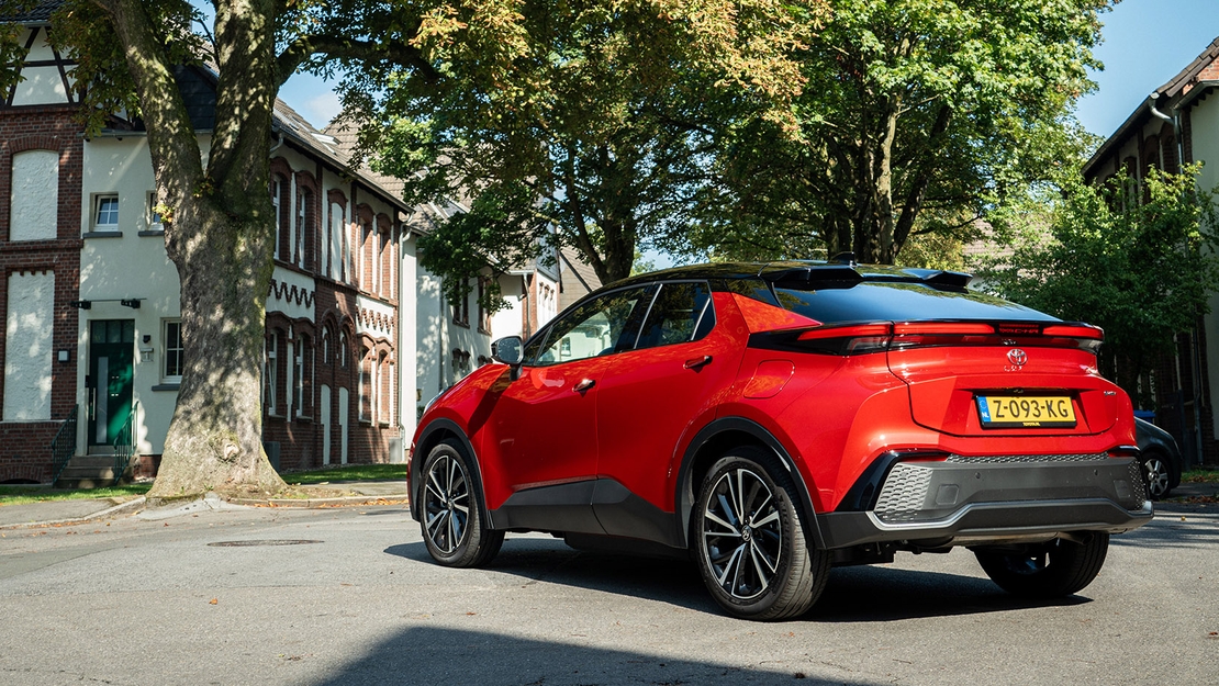 Toyota-C-HR-exterieur-rechter-zijaanzicht-schuin-achter-roodkleurig-voor-bomen-met-huizeng